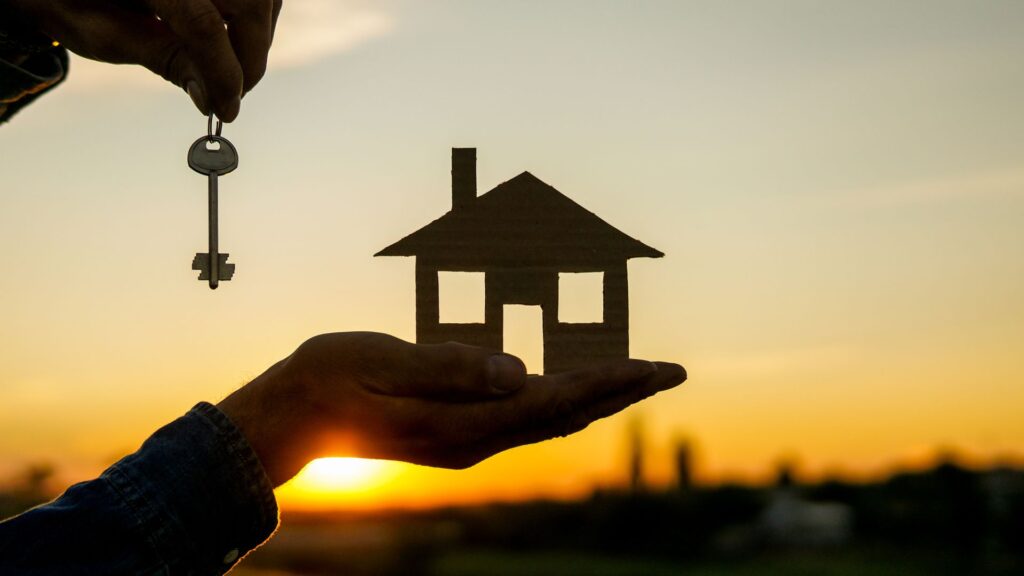 Person holding up a house cutout and a key in a sunset.